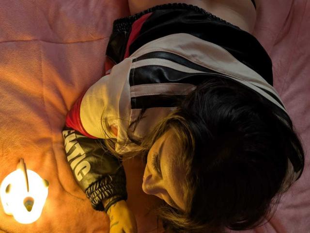 KimyR with dark hair, lying on pink bed, wearing a black and white jacket, red and black shorts, looking down, near a glowing white lamp, face in shadow.