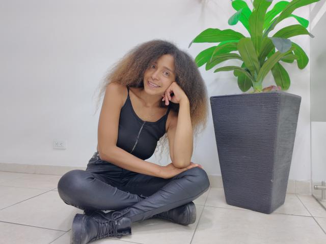 SweetSexx with curly brown hair, black tank top, and black leather pants, sitting cross-legged on tile floor, leaning on hand, smiling, next to green potted plant, wearing black boots.