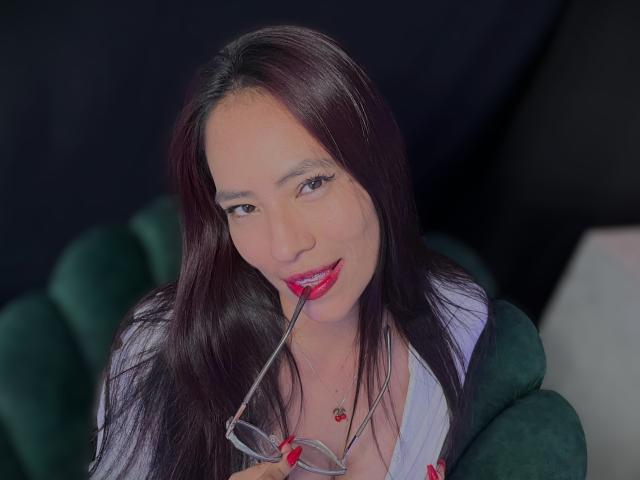SiennaMoretti with long black hair, red lipstick, white shirt, holding glasses in her mouth, green chair, red nails, necklace with red charm, dark background, looking at camera.