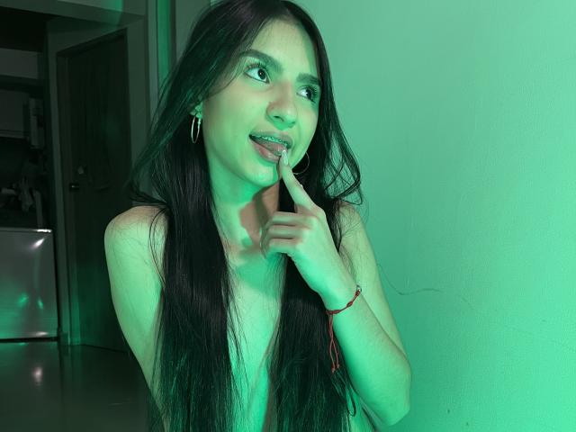 MilanMoon with long dark hair, green-lit face, finger touching tongue, hoop earrings, bracelet, looking sideways, green light reflection, bare shoulders, soft smile, light skin, casual.