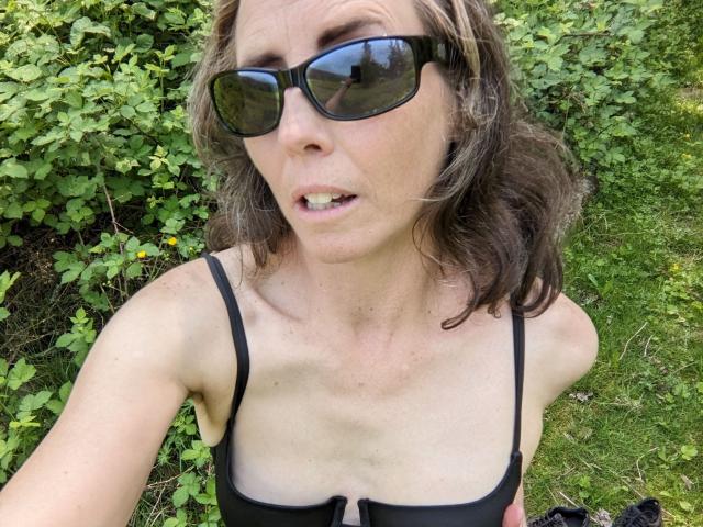 SexyRemmy with medium tits in a black bikini top, wearing black sunglasses, and showing a slight smile. Brown wavy hair, greenery in the background, grass below.