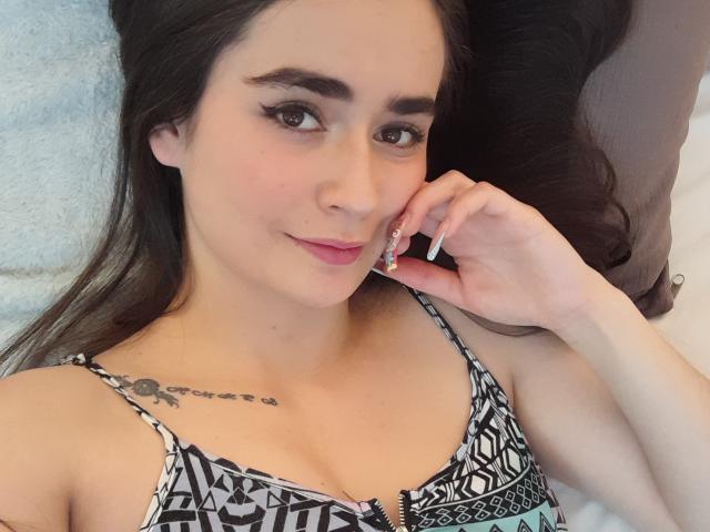 EllieBonneth with dark hair, wearing a patterned top, has small tits, a tattoo on her right shoulder, and is lying down with a slight smile, touching her cheek.