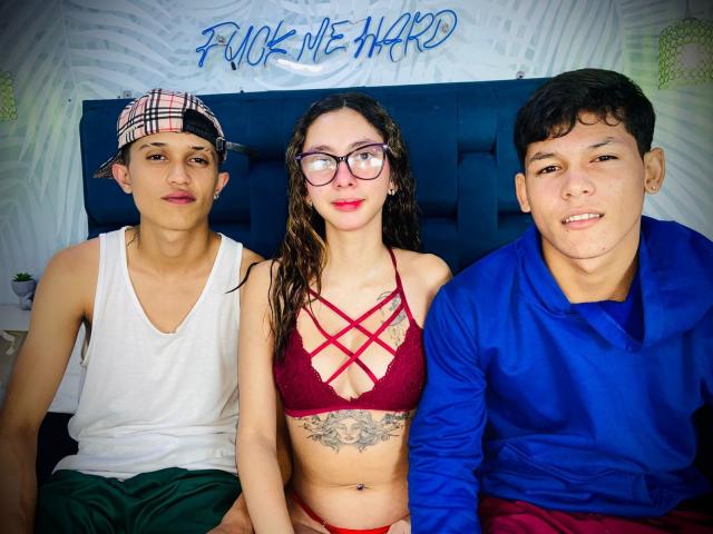 Three people sitting on a bed. HotAndWildy in red lace bra, showing tits, with a tattoo below. HotAndWildy in white tank and green shorts. HotAndWildy in blue shirt.