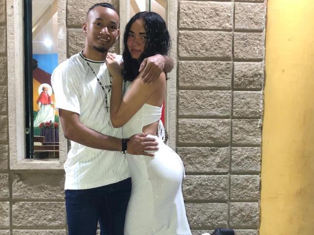CoupleHotxz standing, boy in white tee, black shorts, arms around girl in white cutout dress, hugging her ass, both smiling, brick wall, yellow pillar, window with painting.