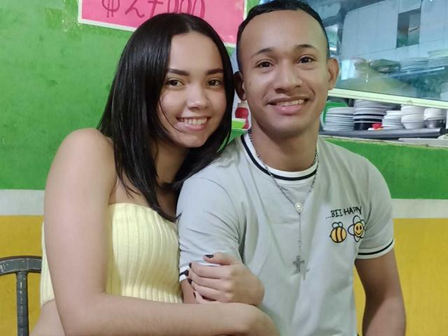 CoupleHotxz with long black hair, yellow strapless top, and CoupleHotxz with short dark hair, gray t-shirt, and cross necklace, smiling and hugging in a green and yellow room.