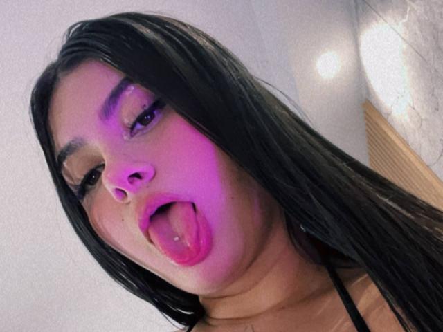 AlanaaWalker with long black hair, purple glow on face, sticking out pink tongue, looking up with half-closed eyes, wearing black top, in selfie.