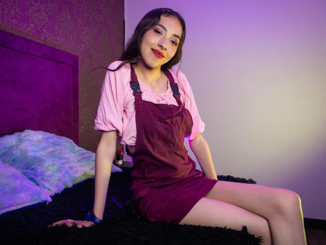 MichelleHollie with red lipstick, brown eyes, and long dark hair, wearing a pink shirt and maroon dress, sitting on a black fuzzy bed, smiling at the camera.