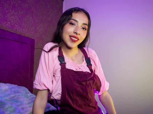 MichelleHollie with dark hair, pink shirt, and burgundy overalls, smiling with red lipstick, purple and pink lighting, bed with purple and blue covers in the background.
