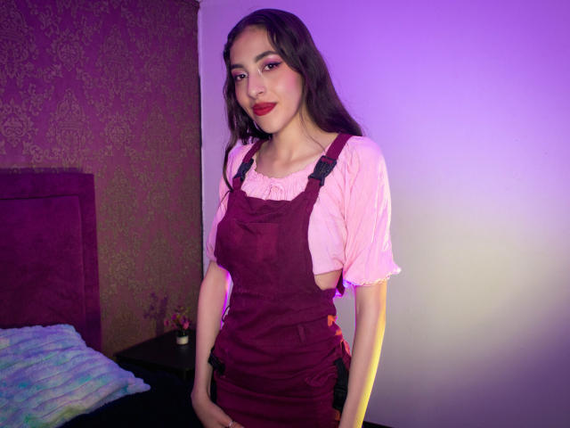 MichelleHollie with long dark hair, red lipstick, pink shirt, and maroon dress, standing, hands in pockets, smiling, bedroom with purple and pink lighting. Boobs visible under dress.