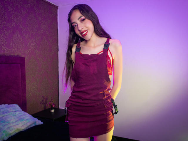MichelleHollie in a red dress, hands behind back, smiling with red lipstick, long brown hair, light skin, purple and pink lighting, bed with blue blanket on the left.