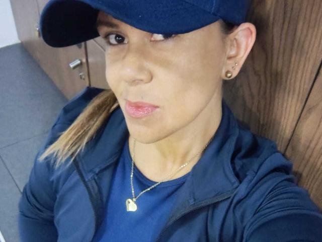 LilithBonneth has a light brown complexion and is wearing a navy blue cap, navy blue shirt, and navy blue jacket. She has a gold heart-shaped necklace and small gold earrings. Her lips are slightly pursed with pink lipstick. She has a small, light brown birthmark above her right eyebrow. Her blonde hair is pulled back in a ponytail. The image is a close-up selfie. The wooden door is in the background.