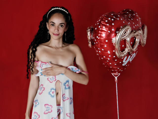 TerryGreen with curly black hair, pearl headband, and earrings, holding a cut-out dress covering her tits, standing next to a red heart balloon against a red background.