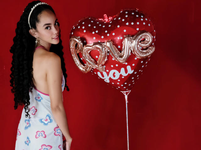 TerryGreen with curly black hair, wearing a white dress with pink and blue hearts, holding a shiny red heart balloon with "love you" and "love" in gold letters.