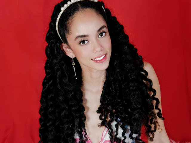 TerryGreen with long, curly black hair, wearing a pearl headband and cross earrings, smiling at the camera. Red background. TerryGreen's tits are covered by a pink and white top.