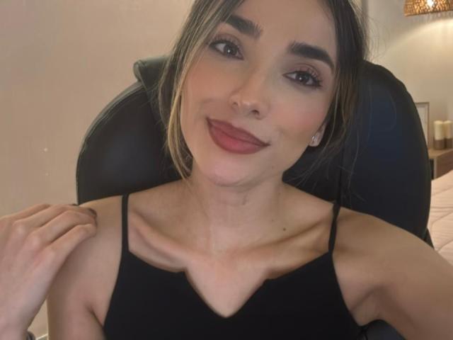AdharaCooperr has a cute smile, brown eyes, and light makeup. She's wearing a black spaghetti-strap top, showing her small tits, and has her hand on her chair.