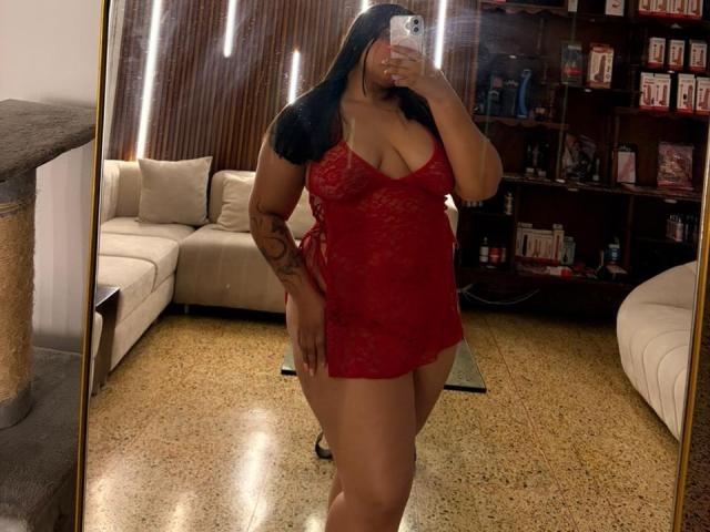 DakotaJhons69 in red lace lingerie, showing her boobs and ass, with long black hair, tattoo on right arm, holding phone, mirror selfie in living room.