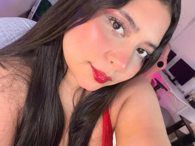 DakotaJhons69 with shiny pink eyeshadow, red lipstick, and long black hair, smiling at the camera. She has a cute face and is wearing a red top.