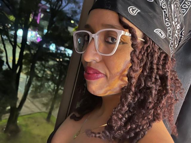 ArtemisaS with curly brown hair, black bandana, white glasses, red lipstick, and a gold necklace, looking slightly to the side. Boobs partially visible, car window in background.