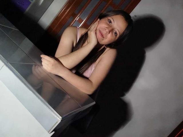 GraceRedMiller with long brown hair, pink tank top, leaning on glass table, hand on cheek, shadow on wall, slight smile, arms crossed, photo.
