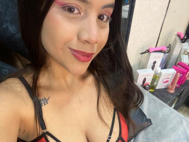 MaddieEvans with dark hair, pink eyeliner, red lipstick, black and red bra, small tattoo on right chest, smiling, seated, visible dildo and pink sex toy on table.