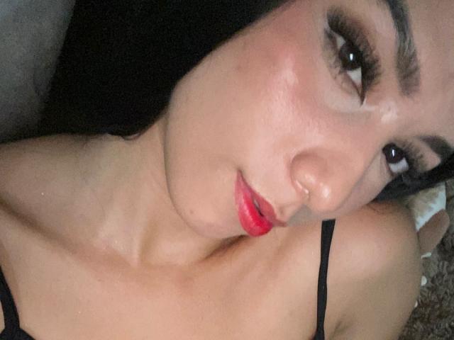 AnieCartier's face with red lips, black eyeliner, and dark hair, looking up with brown eyes, wearing a black strap top, showing her bare collarbone and upper chest.