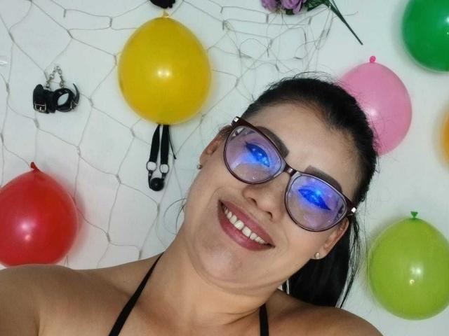 ValeriaLoveer, con cabello negro recogido en una coleta, usando gafas azules reflectantes, sonriendo con lápiz labial rojo y un top halter negro, rodeada de globos de colores y una decoración de telaraña..