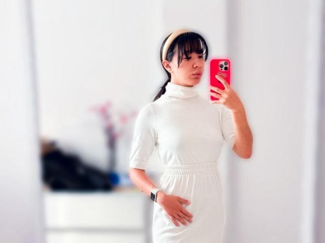 SuriValmont with black hair in a ponytail, wearing a white dress and beige headband, holding a red phone, standing, hand on belly, wearing a black wristwatch.