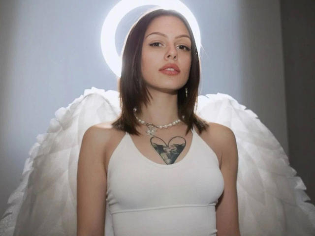 PrettyEllie-hot stands center-frame with a serious expression. She has straight, shoulder-length brown hair and wears a white halter top. Her chest features a black heart tattoo with a small skull inside. She has a pearl necklace with a heart-shaped pendant. Behind her are large, white, feathery angel wings. A bright, circular light halo surrounds her head, highlighting her face. The image is sharply focused, with a neutral, grey background.