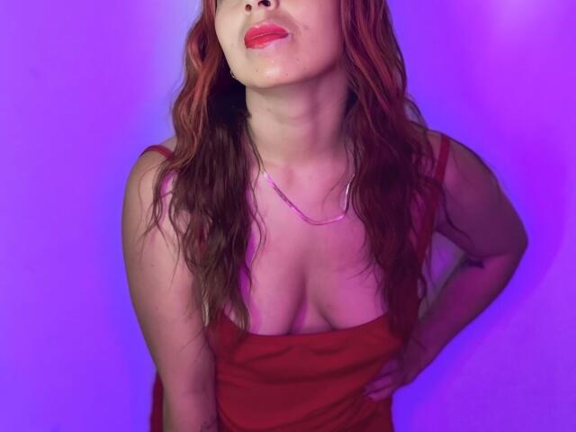 MeghanStorm69, vêtue d'une robe rouge, montrant ses seins, les mains sur les hanches, cheveux roux ondulés, rouge à lèvres rouge, lumière violette, collier, tête légèrement inclinée, confiante, sexy..