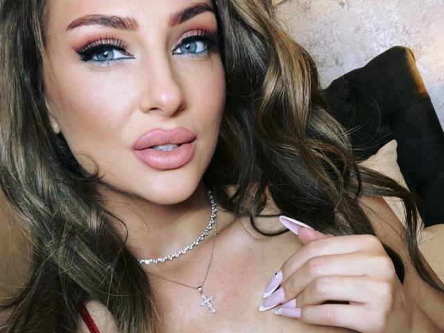 YaseminSaf has blue eyes, full pink lips, and long wavy brown hair. She's wearing a silver necklace with a cross, touching her chest, and showing off her pink nails.