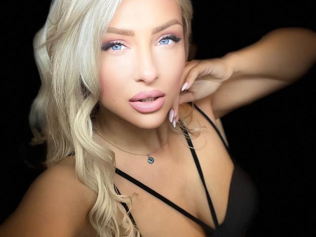 YaseminSaf has blonde wavy hair, blue eyes, pink lipstick, and a black top. She's touching her cheek with a hand, wearing a necklace with a small blue gem.