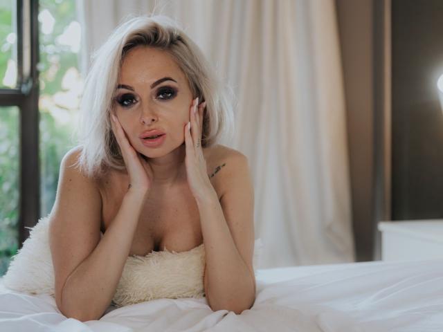 JaquelinMerlo, a blonde woman with shoulder-length hair, leans on a white fluffy pillow, hands resting on her cheeks. She has dark, dramatic eye makeup, a small nose piercing, and a tattoo on her right shoulder. She's wearing a strapless top, exposing her cleavage. The image is softly lit, with natural light coming from a window on the left. Her expression is slightly pouty, and her pink lips are slightly parted. The texture of the pillow contrasts with the smoothness of her skin. The overall composition is centered and focuses on JaquelinMerlo's face. The color palette is mostly neutral with white and beige tones.