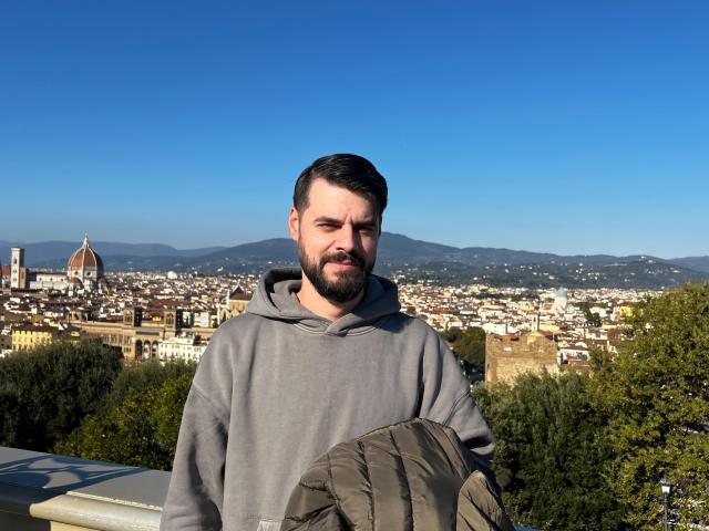 HornyMastter has a beard, short black hair, and wears a gray hoodie, holding a green backpack, standing against a clear blue sky and cityscape.