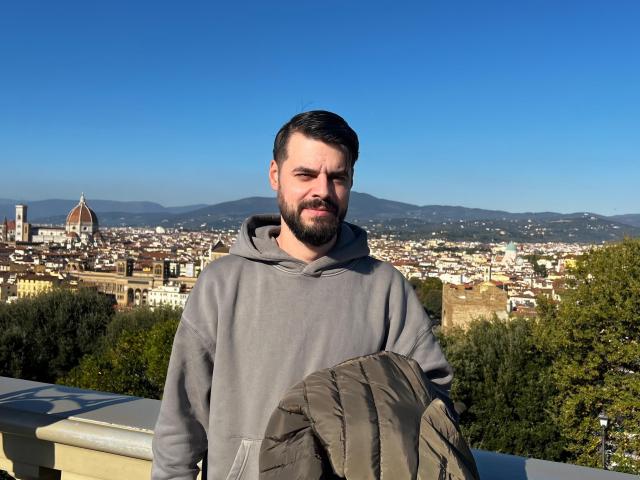 HornyMastter, bearded dude in gray hoodie, holding puffy jacket, standing on balcony, clear blue sky, city with dome in background, mountains in distance, looking slightly annoyed.