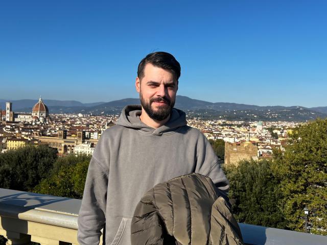 HornyMastter with a beard, gray hoodie, holding a green backpack, standing on a sunny rooftop, blue sky, mountains, and city in the background, smiling slightly.