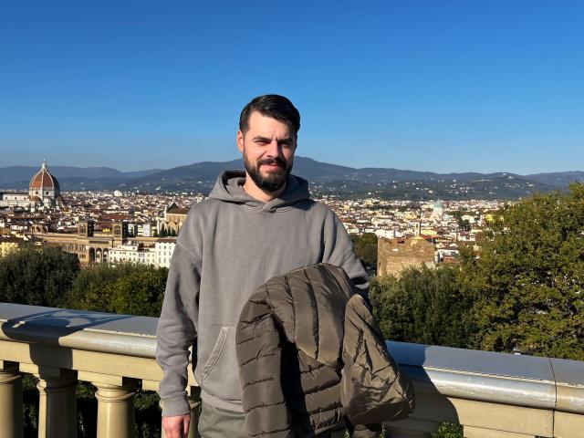 HornyMastter, a bearded dude with short brown hair, wearing a gray hoodie, holding a green jacket, standing in front of a balcony with a city and mountains in the background.