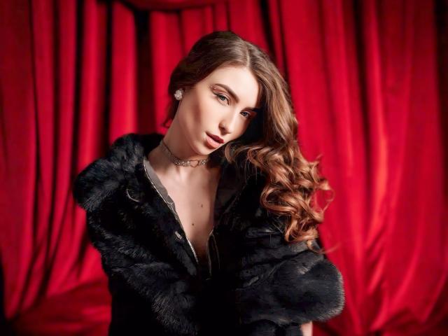 MelanieFoster with wavy brown hair, wearing a black fur jacket, choker, and earrings, looking seductively at the camera. Red velvet curtains in the background.