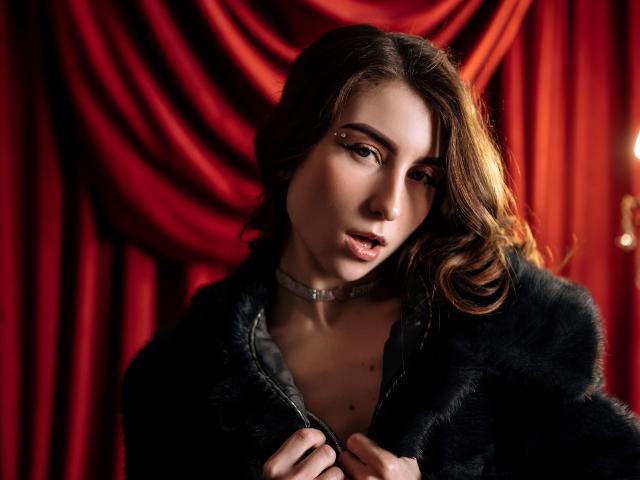 MelanieFoster with dark wavy hair, wearing a black fur coat and silver choker, holding it open to reveal her small tits, against a red curtain.
