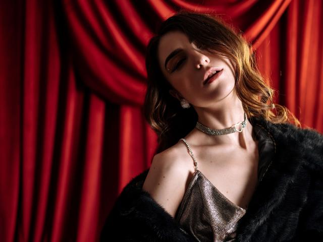 MelanieFoster has wavy brown hair, eyes closed, and slightly parted lips. She wears a sparkling silver dress with thin straps and a black fur coat. A glittering choker necklace and diamond earrings are visible. Her head is tilted back, and her expression is relaxed. The red velvet background is draped with soft, flowing folds. The lighting highlights her facial features and the texture of the fur coat. The composition is intimate and luxurious.