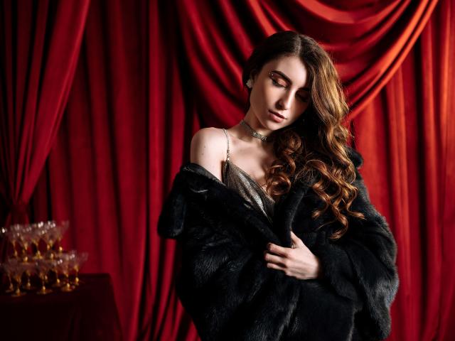 MelanieFoster with long, wavy brown hair, wearing a black fur coat, silver dress, and choker, looking down, holding coat, red curtains, and gold champagne glasses in background.