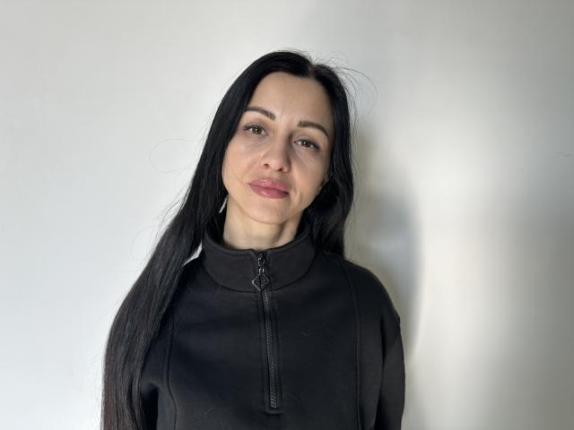 EvaClarkX, com cabelos longos e pretos, vestindo uma camisa preta com zíper, tem um sorriso discreto, seios médios, pele clara e olhos castanhos, e está em pé em frente a uma parede branca lisa..