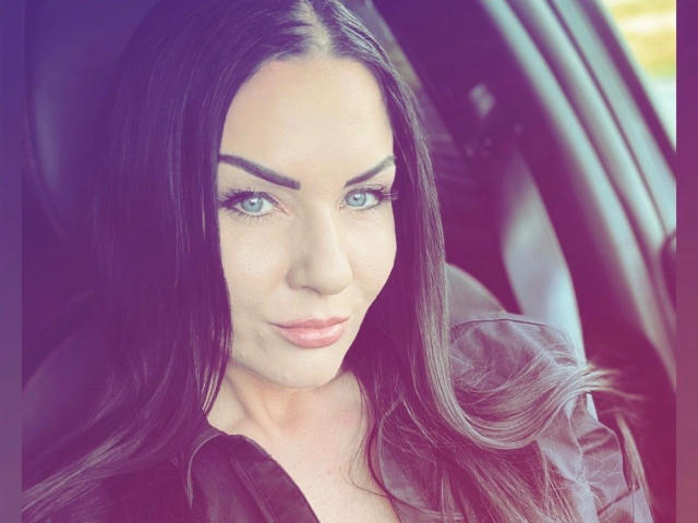 Close-up of Malina-Lay with long, straight black hair and green eyes. She has a serious expression with slightly parted lips. Her eyebrows are thick and arched. She's wearing a dark, sheer top with a subtle collar. The light is soft, highlighting her facial features. The image has a slight purple tint. Malina-Lay's skin is fair, and her makeup is subtle with defined eyeliner and light pink lipstick. The background is blurred, keeping focus on her face.