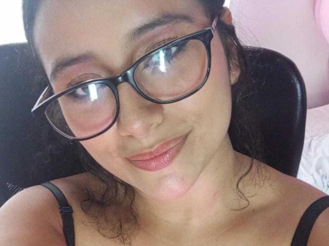 NicollWest with glasses, brown eyes, and a slight smile, showing her shiny pouty ass-adjacent lips. She's wearing a black strap top, and has wavy dark hair.