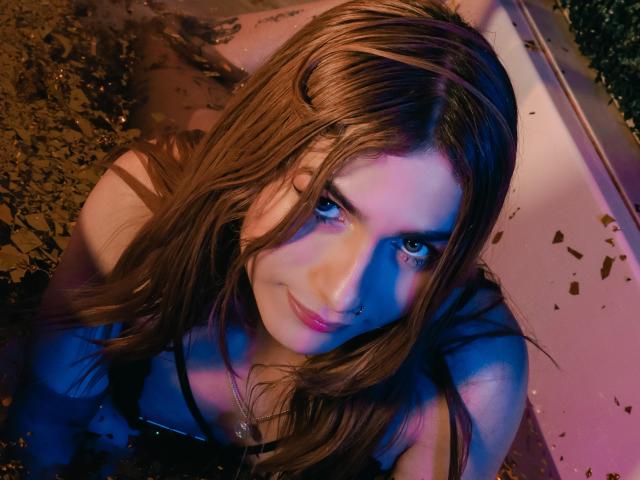 AndyDupont, a young woman with wet, medium-length brown hair, leans forward in a shallow, muddy bath. Her skin is fair, and she has a subtle, confident smile. She wears a black strap, and her nose is pierced with a small hoop. The image is illuminated with blue and pink lighting, creating a moody, vibrant effect. Her eyes are focused on the camera, and her hair is slightly tousled. The muddy water has small brown particles. The composition is tight, emphasizing her face and upper shoulders.