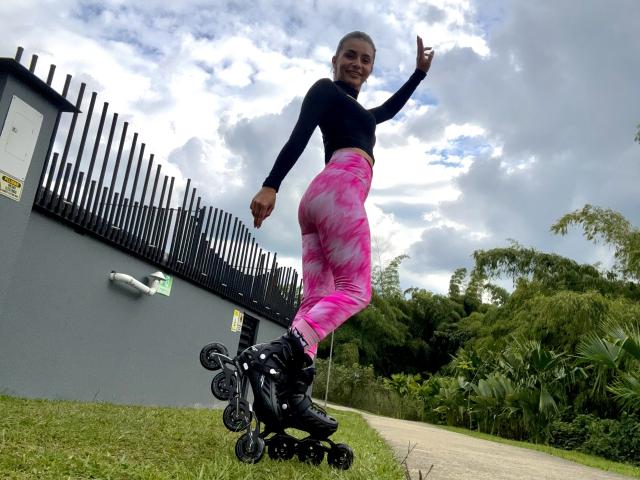 KimmyK, con una figura snella e atletica, indossa una maglia a maniche lunghe nera e leggings rosa tie-dye, è in piedi su un paio di pattini a rotelle neri, punta verso l'alto, con erba verde e alberi sullo sfondo..