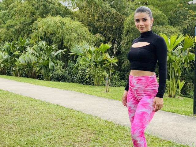KimmyK has pink tie-dye pants, black long-sleeve crop top with a cutout, and is standing on a grassy path with a smile, looking at the camera.