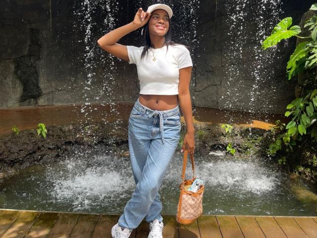 HazelClark with dark skin, black hair, white cap, white crop top, blue jeans, white sneakers, holding tan bag, standing under water spray, smiling, showing her small tits.