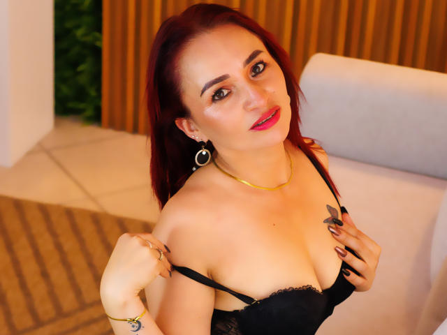 EmaCollins with red hair, black lace bra, and gold necklace, pulling straps down, showing her tits, red lipstick, and ringed fingers, smiling at the camera.