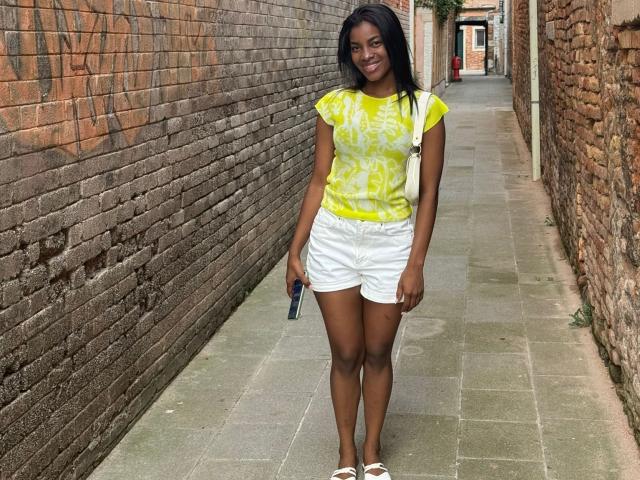 HazelClark has dark skin, long black hair, wearing a yellow tie-dye shirt, white shorts, white sneakers, and a white strap bag, standing in a brick alleyway.