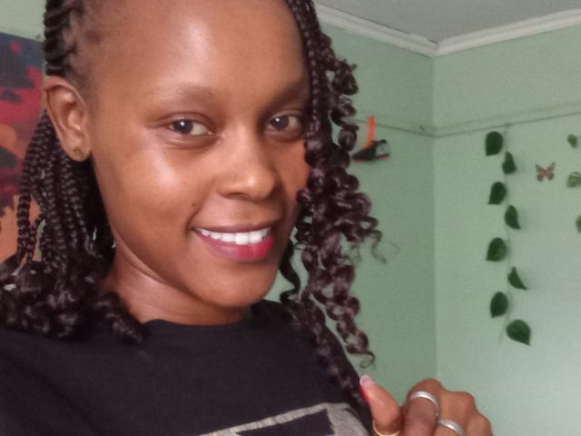IvyTheeGodess with dark brown skin, curly black hair, and pink lipstick, smiling at the camera, wearing a black and gray shirt, showing a ring on her right hand.